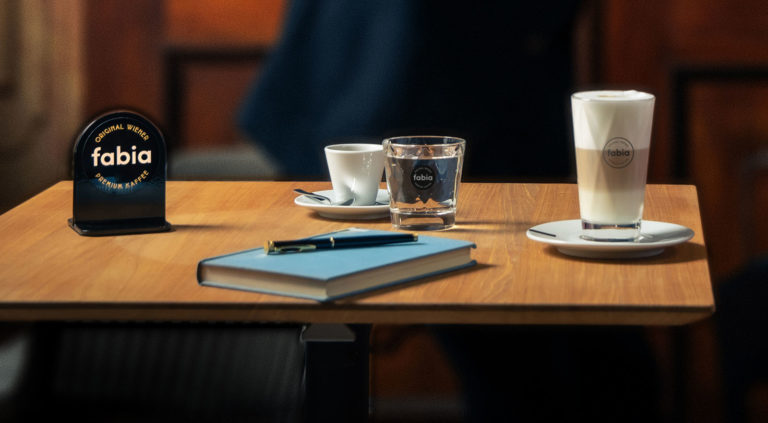Ein Kaffee Latte, eine kleine Tasse Kaffee, ein Glas Wasser und ein Notizbuch auf einem Tisch liegend frontal fotografiert.