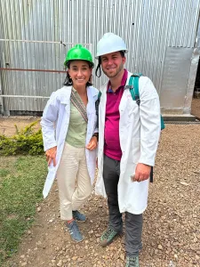 Anna und Lukas lächelnd vor der Produktionsstätte in Tansania. Sie tragen einen Helm und einen weißen Mantel.