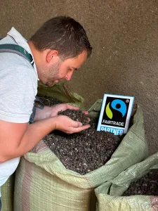 Lukas hat einen Haufen Kaffee Bohnen in seiner Hand und reicht daran. Vor ihm steht ein Sack Faitrade Kaffee